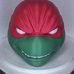 May include: A 3D-printed mask of a Teenage Mutant Ninja Turtle character. The mask features a red head covering, white eyes, and a green face with a wide grin. The mask is sitting on a white surface.