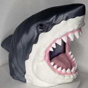 Op de afbeelding: Een haaienhoofdsculptuur met een open mond, met scherpe witte tanden en een roze tong. Het lichaam van de haai is donkergrijs, met witte en roze accenten. De sculptuur is een decoratief item.