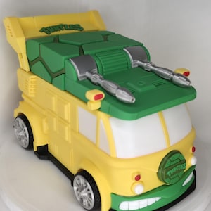 May include: A yellow and green toy van with the word "Turtles" on the spoiler. The van has silver-colored details, including the wheels and two side-mounted weapons. The front of the van has a green bumper with a smiling mouth design.