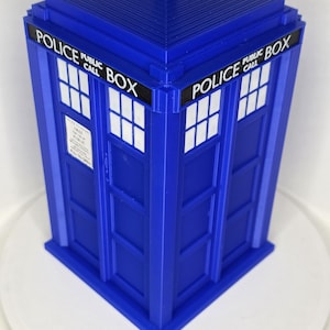 May include: A blue miniature replica of a British police box, with the text "POLICE PUBLIC CALL BOX" on the top and sides. The model has white window panes and a small roof detail.