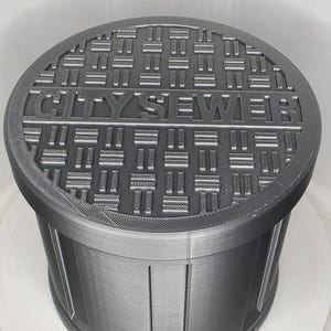 May include: A dark gray cylindrical container with a circular lid featuring a raised grid pattern and the text CITY SEWER embossed across the center. The sides of the container have vertical ridges and a smooth finish, mimicking a manhole cover.