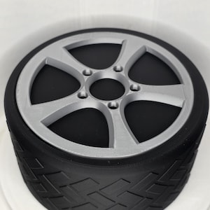 May include: A black tire with a textured tread pattern and a silver five-spoke wheel rim. The wheel features a central hub with five circular bolt holes arranged in a symmetrical pattern, contrasting against the matte black rubber-like tire surface.