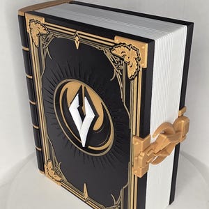 May include: A black book with gold accents and a white emblem. The book has a gold clasp and decorative corner pieces. The pages are white, and the cover features a detailed design with a central emblem.
