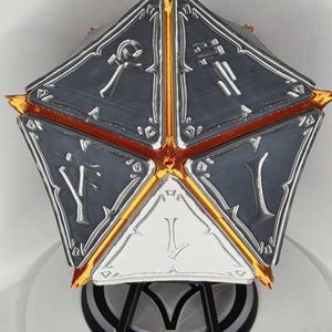 Op de afbeelding: Een veelzijdig, geometrisch object met een vijfhoekige vorm. Het object is voornamelijk grijs met zilveren en gouden accenten. Elke kant heeft geëtste symbolen. De randen zijn goud en rood omlijnd. Het object wordt gepresenteerd op een wit oppervlak.