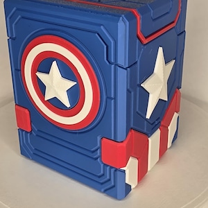 May include: A blue, red, and white rectangular storage box featuring a raised Captain America shield emblem on the front and a white star on the side. The top surface displays a white letter A centered within a geometric pattern. The design incorporates red and white stripes along the bottom edge, with a textured finish on the blue sections and a smooth appearance on the white elements.