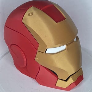 Op de afbeelding: Een rood en goudkleurige helm met een glad, gesegmenteerd ontwerp, een centrale voorplaat, een opvallend voorhoofdgedeelte en witte oogopeningen. De helm heeft een afgeronde bovenkant, gebogen zijpanelen en een kinbeschermer in matte kleuren.