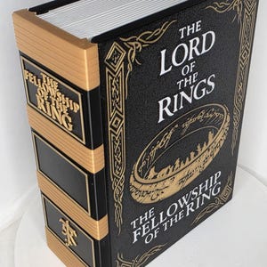 Puede incluir: Una caja decorativa en forma de libro negro y dorado titulada The Lord of the Rings: The Fellowship of the Ring. La portada presenta un gráfico de anillo dorado con escritura élfica, mientras que el lomo muestra el título y un símbolo dorado.