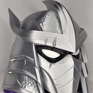 May include: A silver and grey Shredder helmet, with sharp, angular details and a prominent crest. The helmet has a metallic sheen, with black eye coverings and a purple base. The design is reminiscent of the Teenage Mutant Ninja Turtles.
