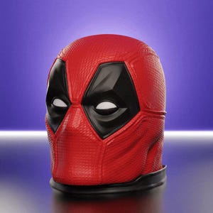 May include: A red Deadpool mask with black eye and mouth details. The mask has a textured surface and a black base, set against a purple and blue gradient background. The mask is a replica of the character's iconic look.