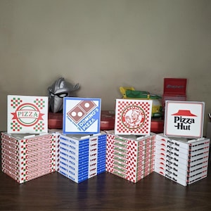 May include: A collection of miniature pizza boxes from various pizza chains, including Pizza Hut, Domino's, and Antonio's. The boxes are stacked and arranged on a wooden surface, with the logos and branding clearly visible.