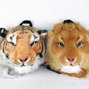 Op de afbeelding: Twee rugzakken in de vorm van dierenkoppen van pluche: een tijger met oranje en zwarte strepen en een leeuw met een bruine manen. Beide hebben realistische kenmerken, waaronder gouden ogen en roze neuzen. De rugzakken hebben zwarte banden.
