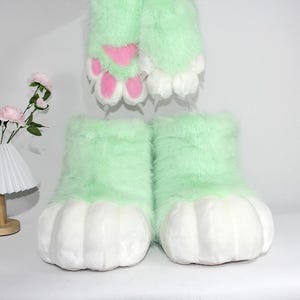 May include: A pair of light green furry costume feet with large white paw pads. The top of the feet are a soft, light green material. The paw pads are white with pink accents. A matching pair of furry hand paws are visible in the background.