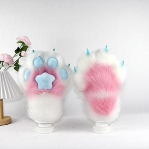 May include: Two plush paw-shaped gloves. The gloves are white with pink and blue accents. The paw pads are pink with a blue star in the centre. The claws are blue. The gloves are displayed on white stands.