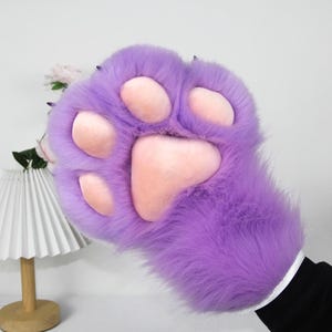 May include: A large, plush purple paw with pink pads and black claws. The furry paw is detailed with a realistic design, and the wrist is covered with a white band and black sleeve.