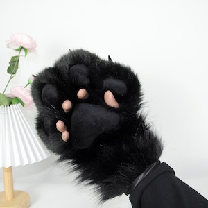 Könnte beinhalten: Ein schwarzer Pfotenhandschuh aus Kunstfell mit realistischer Gestaltung. Der Handschuh hat schwarzes Fell, ein schwarzes Pfotenpolster und rosafarbene Krallen und Pfotenpolster. Der Handschuh ist so konzipiert, dass er über die Hand und das Handgelenk passt.