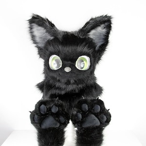 May include: A plush creature head with long, pointed black fur ears, thick black faux fur, and large, round, light green eyes. The creature features a small grey nose and two large, black, padded paws positioned in the foreground.