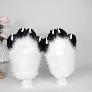 May include: A pair of plush paw gloves with black tops and white fur. Each paw has three white claws. The gloves are displayed on white cylindrical stands against a white background. The gloves are a cosplay accessory.