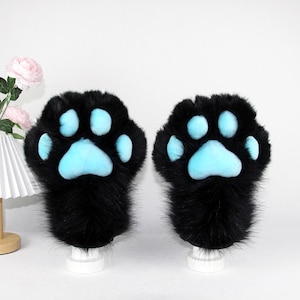 May include: A pair of black faux fur paw gloves with light blue paw pads. The gloves are designed to resemble cat paws, with a fluffy texture and a playful aesthetic. The gloves are displayed on white stands.