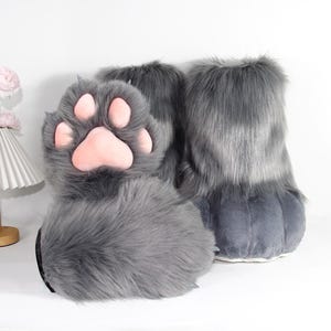 May include: A pair of gray faux fur boots designed to resemble animal paws. The boots feature long, shaggy gray synthetic fur, with a prominent soft pink paw pad on the front of each boot, complete with four rounded toe pads and one large heel pad.