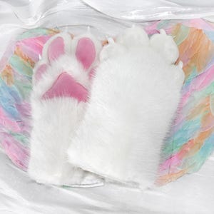May include: A pair of white plush paw gloves with pink paw pads. The gloves are fluffy and soft, with a realistic design. The background features a colorful, feathered wing.