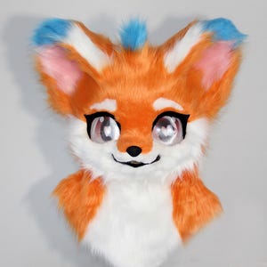 May include: A vibrant orange, white, and blue fox fursuit head with large, expressive eyes. The fox has a friendly expression, with a black nose and a smiling mouth. The ears are tipped with blue, and the inside of the ears are pink.