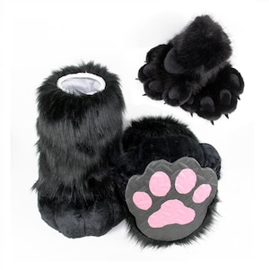 May include: A pair of black faux fur animal paw slippers featuring long black synthetic fur, a white interior lining, and a dark gray sole with five distinct pink paw pads. The slippers are designed to resemble large, plush animal feet.