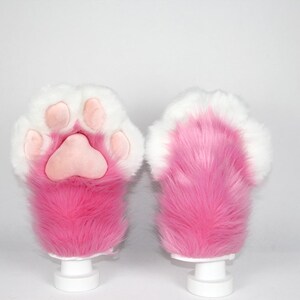 May include: Pair of pink and white faux fur paw gloves. The gloves have a pink paw pad and pink fur with white fur around the edges. The gloves are displayed on white stands against a white background.