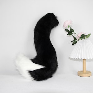 Puede incluir: Una cola de piel sintética negra y blanca, con forma curvada. La cola es principalmente negra, con una transición a blanco en la punta. La cola es esponjosa y parece suave. El fondo es blanco.