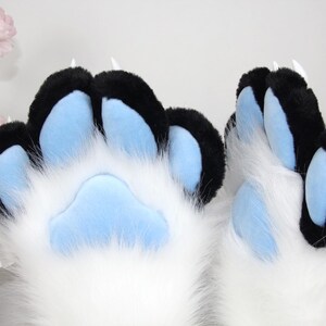 May include: Close-up of furry paw pads in white, black, and light blue. The paw pads have black tips and light blue pads. The fur is white and fluffy. The claws are white and pointed.