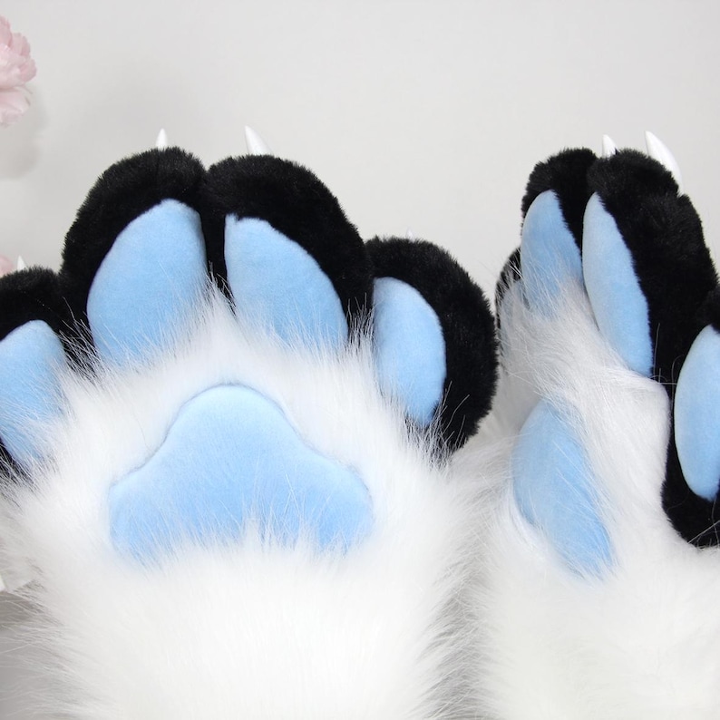 May include: Close-up of furry paw pads in white, black, and light blue. The paw pads have black tips and light blue pads. The fur is white and fluffy. The claws are white and pointed.