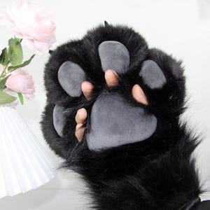 May include: A black furry paw with gray paw pads and visible claws. The paw is made of soft, plush material. The image shows a close-up of the paw, with a person's fingers visible inside.