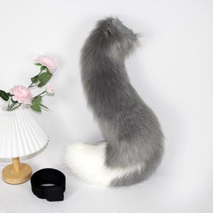 May include: A fluffy, gray and white faux fur tail. The tail is long and curved, with a gray body and a white tip. A black belt with a buckle is at the base of the tail. Decorative pink roses and a white lamp are in the background.