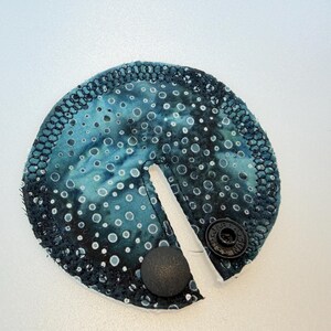 Op de afbeelding: Een rond, teal stoffen item met een patroon van witte en lichtblauwe cirkels. De rand is afgewerkt met een zwarte, getextureerde rand. Het heeft een spleet en twee zwarte druksluitingen. Er is ook een zwarte, ronde knop bevestigd.