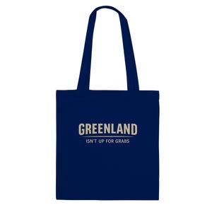 May include: A navy blue tote bag featuring the word "GREENLAND" in a light tan font, with the text "ISN'T UP FOR GRABS" underneath. The bag has two navy blue handles.