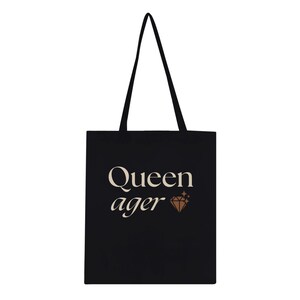 May include: Black tote bag featuring the text "Queen ager" in an off-white font, accompanied by a brown diamond graphic. The bag has long black handles and is made of a durable fabric, ideal for carrying items.