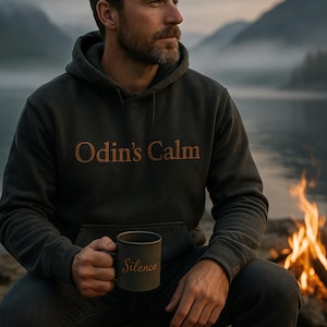 May include: A dark grey hoodie with the text "Odin's Calm" in orange lettering. The person is holding a dark grey mug with the word "Silence" printed on it. A second mug with the same text is on a rock. The scene is set outdoors with a fire.