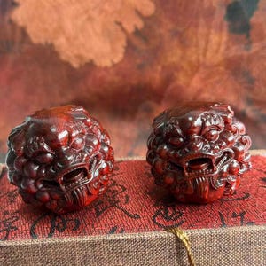 May include: Two intricately carved, reddish-brown wooden sculptures of mythical creatures. Each sculpture features detailed facial features and a textured, spherical body. The carvings are displayed on a red patterned surface.