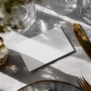 May include: A mock-up scene featuring a white card, crystal glassware, and gold cutlery on a white linen tablecloth. The scene is illuminated by natural light, creating shadows. The text "PEARLY PAPER MOCKUPS" is visible.
