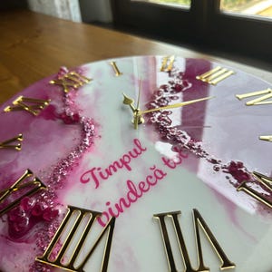 Può includere: Un orologio da parete rotondo con una superficie in resina marmorizzata bianca e magenta, caratterizzato da numeri romani dorati e lancette dorate. Una fascia diagonale di cristalli magenta frantumati attraversa il centro, con il testo "Timpul vindecă tot" in corsivo magenta.