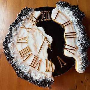 Può includere: Un orologio da parete rotondo con quadrante in resina marmorizzata bianca e nera e numeri romani dorati. I bordi sono decorati con texture cristalline bianche e nere. L'orologio ha lancette dorate e un design diviso.