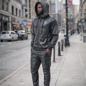 Può includere: Una tuta nera e argento con un motivo geometrico. La tuta include una giacca con cappuccio e zip e pantaloni abbinati. La persona nell'immagine è in piedi su un marciapiede della città.