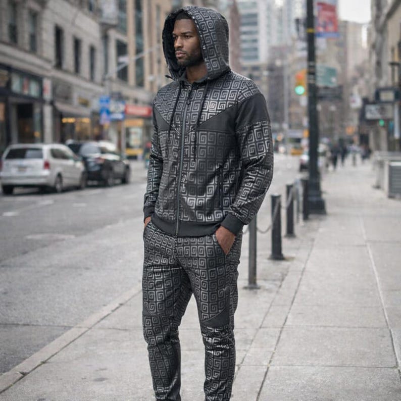 May include: A black and silver tracksuit with a geometric pattern. The tracksuit features a hooded zip-up jacket and matching trousers. The person in the image is standing on a city pavement.