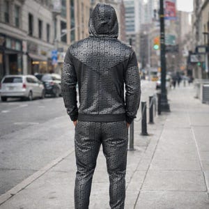 May include: A black tracksuit with a geometric pattern, including a hooded jacket and matching trousers. The jacket has a zip closure and the pattern is a repeating square design. The person is standing on a pavement in a city.
