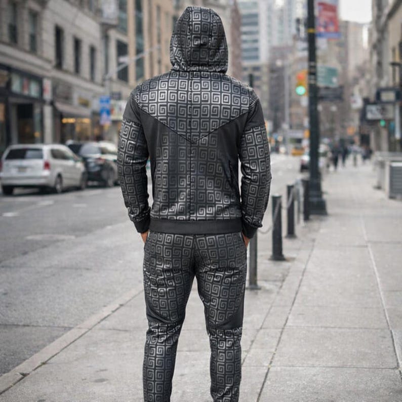 May include: A black tracksuit with a geometric pattern, including a hooded jacket and matching trousers. The jacket has a zip closure and the pattern is a repeating square design. The person is standing on a pavement in a city.