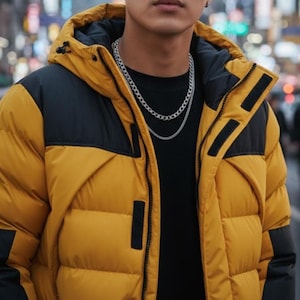 May include: A mustard yellow and black color-blocked puffer jacket with a hood, featuring a black interior lining and a front zipper closure. The jacket is worn over a plain black shirt, complemented by two layered silver-toned chain necklaces.