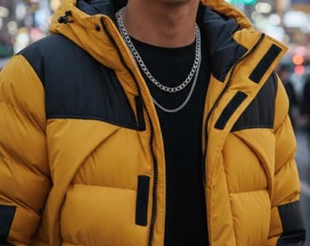 May include: A mustard yellow and black color-blocked puffer jacket with a hood, featuring a black interior lining and a front zipper closure. The jacket is worn over a plain black shirt, complemented by two layered silver-toned chain necklaces.