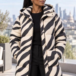 May include: A long-sleeved puffer jacket featuring a black and cream zebra stripe pattern. The jacket includes a hood, a full-length front zipper, and snap button closures. It is worn over a black top and black trousers, providing a layered look.