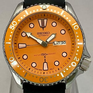 May include: A stainless steel Seiko automatic watch with an orange dial and bezel. The watch features white hands and markers, a black leather strap, and the date displayed at the 3 o'clock position.