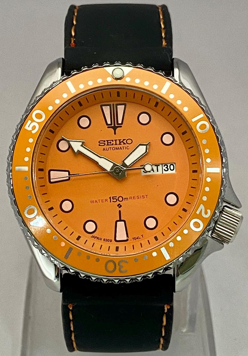 May include: A stainless steel Seiko automatic watch with an orange dial and bezel. The watch features white hands and markers, a black leather strap, and the date displayed at the 3 o'clock position.