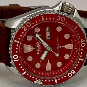 K&ouml;nnte beinhalten: Seiko Automatik-Armbanduhr mit rotem Zifferblatt und braunem Kautschukarmband. Die Uhr verf&uuml;gt &uuml;ber eine silberne L&uuml;nette mit wei&szlig;en Markierungen und eine Datumsanzeige. Das Zifferblatt hat wei&szlig;e Zeiger und Markierungen.
